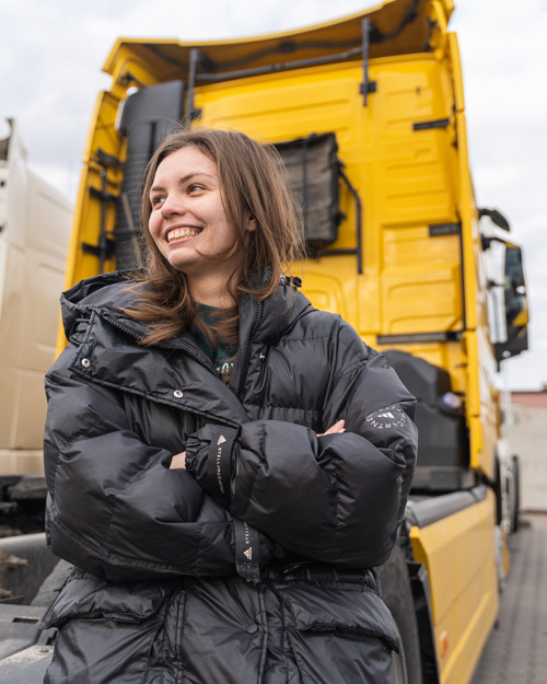 smiling woman in black puffer jacket standing in front of yellow truck showcasing friendship and adventure in transportation industry